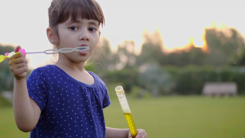 A close-up beautiful happy shot of a cute and adorable little girl child is blowing soap water bubbles against the sun in the park or garden. 