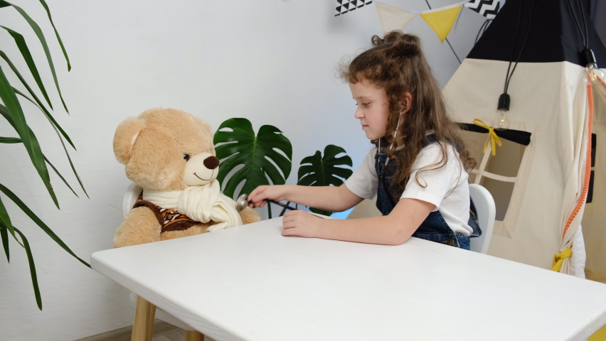 Paediatrics, playtime concept. Little girl kid play doctor with plush bear sit at table near tent. Caring child hold stethoscope touch toy have fun pretending nurse treating fluffy patient at home Royalty-Free Stock Footage #1082941036