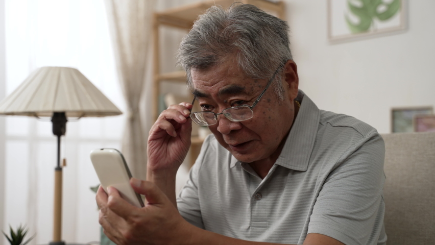 Elderly asian male having eyesight problems and wearing eyeglasses while gazing and reading message on smartphone on sofa. Old man having eye blurred vision indoors and moving glasses up and down Royalty-Free Stock Footage #1082945572