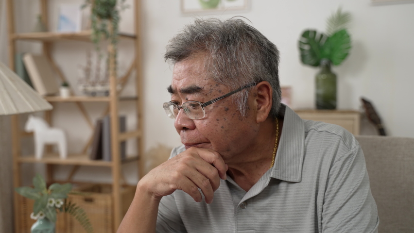Senior asian man deep in thought while sitting on couch in living room at home. serious old male with hands putting under chin and feeling worried on sofa. sad people in glasses with upset emotion Royalty-Free Stock Footage #1082945605