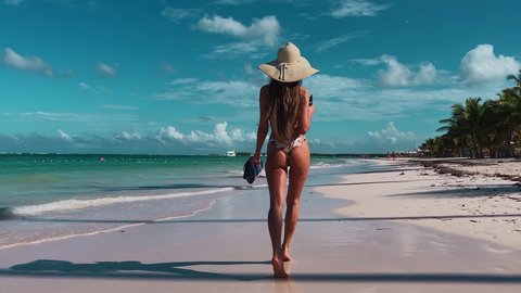 A girl with a beautiful ass is walking along the beach in a swimsuit. A Big Hat is worn on his head. Models, beach, tropics, Dominican Republic, coast, sea, ocean, sand, bikini, tide, palm trees, hat