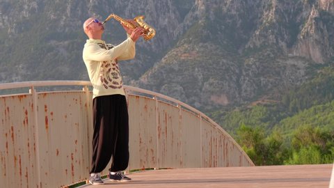 video Young bald man street musician saxophonist in sunglasses stands on bridge and plays golden alt saxophone on musical instrument against background of green mountains, blue sky, sunrise, sunset.