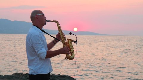 video Young bald man saxophonist in white shirt plays golden alt saxophone on musical instrument, on seashore, against background of sea, beautiful sunset on of Cyprus. Record sound to microphone.