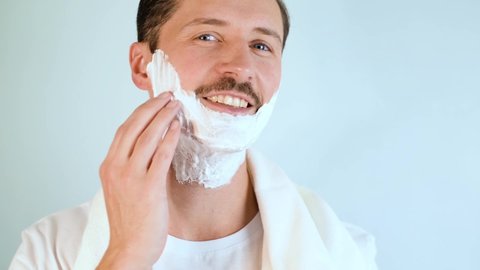 Handsome smiling unshaven young man applying moisturizing shaving foam on face and looking at camera. Portrait of happy man with towel on shoulders performing daily self-care, beauty routine indoors