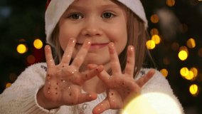 Close up of little girl in red santa hat looks at camera, smiles and shows palms with gold glitters and shiny confetti on background of Christmas tree. Happy New year - Powered by Shutterstock - Get 15% off with code: PIKWIZARD15