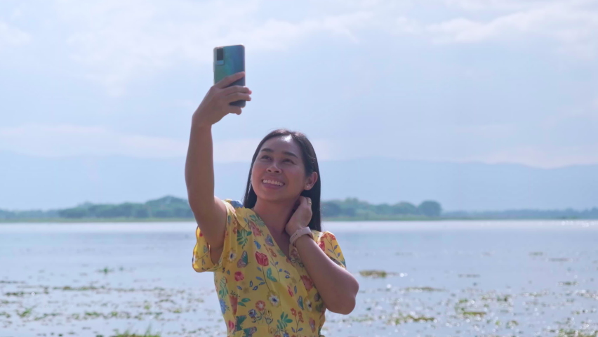 Asian female tourist is using smartphone to take selfie and enjoy the view with happy mood on a sunny day. Outdoor travel and Nature theme.