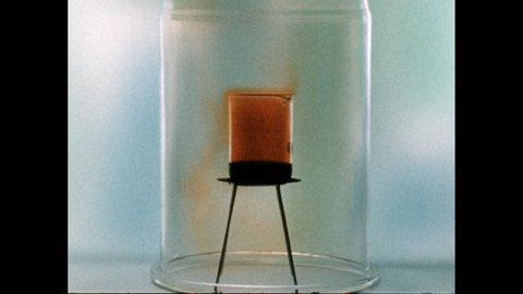 1960s: Gas seeps from flask under glass bell jar. Hand with stick points to molecule models. Gas seeps from flask under glass bell jar. Metal cube sits on plate.