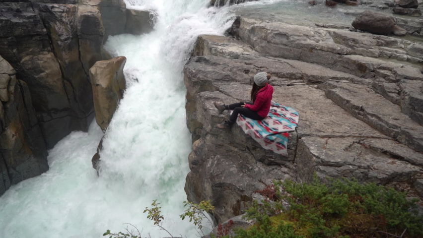Female Sitting on Rock Above Waterfall and Glacial River in Canadian Countryside on Cold Autumn Day. Sunwapta Falls, Jasper National Park, Canada