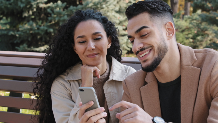 Bearded guy with attractive girl in autumn park sitting on bench cheerful couple interestedly looks at smartphone screen young man actively communicate with girlfriend partners discuss online shopping | Shutterstock HD Video #1082964622