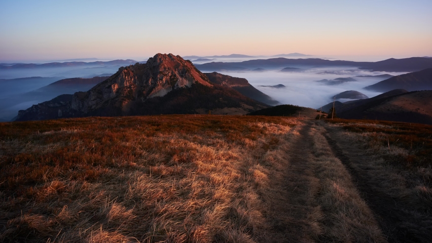 Grassy hiking trail in the mountains. The rocky peak of the mountain is surrounded by low inverse clouds. Pleasant warm morning light at sunrise.
