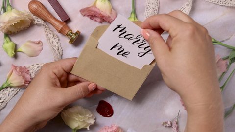 Hands opening and closing envelope with a MARRY ME card over a marble table near pink flowers close up 