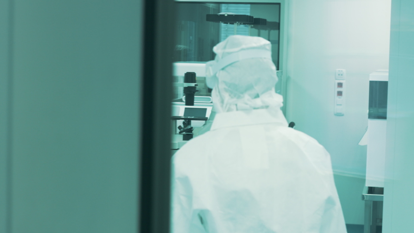 Handheld shot of a lab worker, in a protective suit, passing through a cleanroom stainless steel automatic door with a half view window and entry lights. A scientist working in a laminar flow cabinet. | Shutterstock HD Video #1082970178