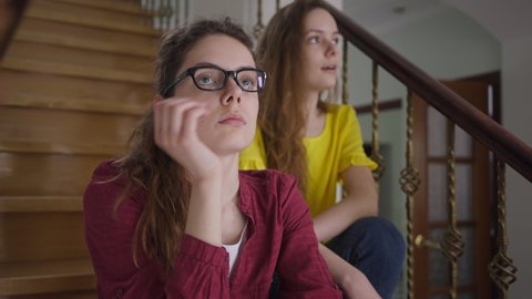 Portrait of bored Caucasian young nerd woman in eyeglasses listening twin sister talking at background gesturing bla-bla. Disinterested beautiful slim millennial lady sitting on stairs with sibling