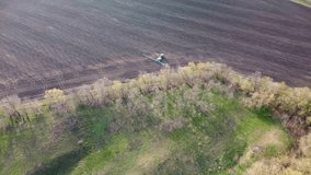 Aerial footage of blue tractor with long booms turning around near green trees and spraying on cultivated land field. Agricultural agronomy spring equipment
