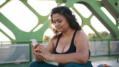 Young overweight woman in sports clothes sitting and using smartphone outdoors in bridge.