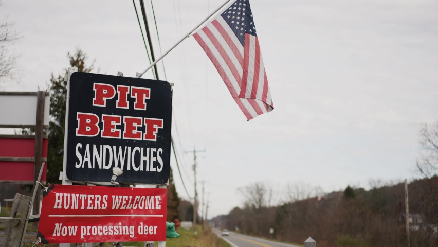 Traffic passing on rural road with Pit Beef and Deer Processing banner signs under US flag.