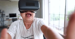 Asian old female sitting on sofa in living room wears vr virtual reality eyeglasses holding joystick while playing game with fun emotion. Concept of learning new technology for senior people.