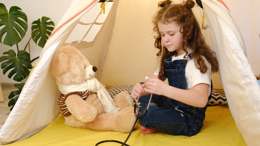 Healthy happy little school girl playing game as vet doctor concept holding stethoscope listening sick toy at home, cute funny child sit in cozy tent pretending medical nurse treat teddy bear patient  Royalty-Free Stock Footage #1082983780