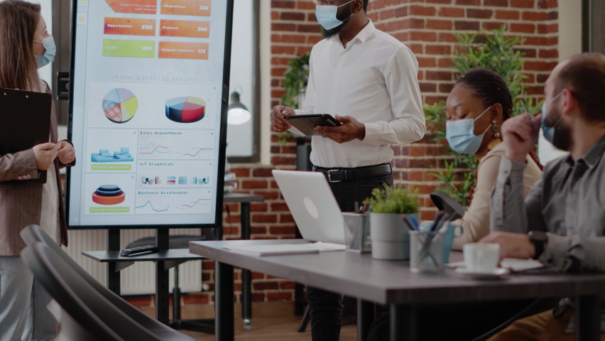 Man and woman with face mask planning business growth in front of coworkers at briefing meeting. Colleagues using monitor to do financial presentation for marketing strategy during pandemic. - Powered by Shutterstock - Get 15% off with code: PIKWIZARD15