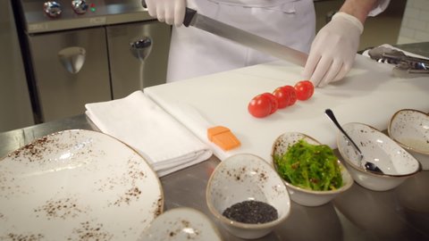 professional chef prepares food in the kitchen. close-up male hands in rubber gloves cut a red tomato on a white surface with a long knife, dolly shot. work in the restaurant kitchen. copy space