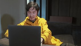 An Asian guy sits on sofa in a small office and communicates via video link through a laptop
