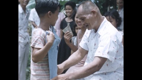 1950s: Party. Little boy kisses old man's hand. Boy unwraps gift. Man holds up shirt to boy. Hand holds shooting slate. People pack rice into bowls.