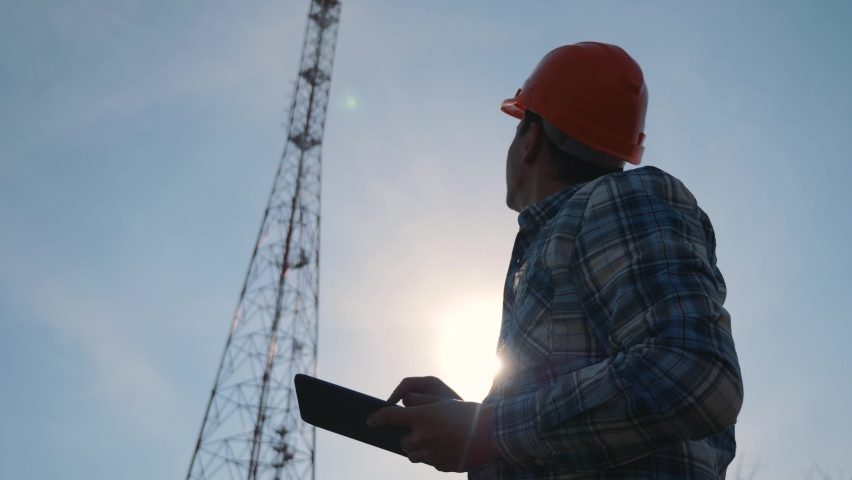 A silhouette of a telecommunications network engineer in a hard hat uses a tablet at work. Royalty-Free Stock Footage #1083006421