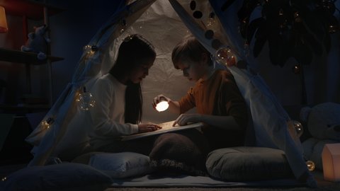 Teens in decorative makeshift hut looking at book page in evening. Young boy and girl using flashlight while sitting on floor and spending free time together. Concept of leisure