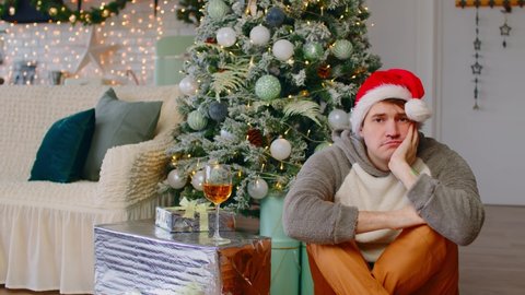 Sad man in santa hat sitting on floor near Christmas tree in living room. Lonely male sighs, looking at camera then down during celebrating holidays.