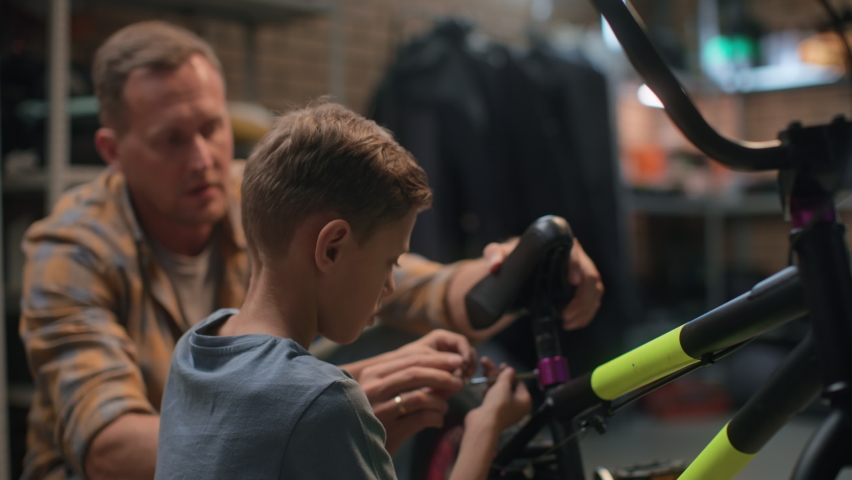 Repair bicycle by small boy in garage. Father helps attentive son to fix bike. Maintenance care and installation of bicycle. Dad explains to young cycle repairman indoors. Happy fatherhood and grow up Royalty-Free Stock Footage #1083013060