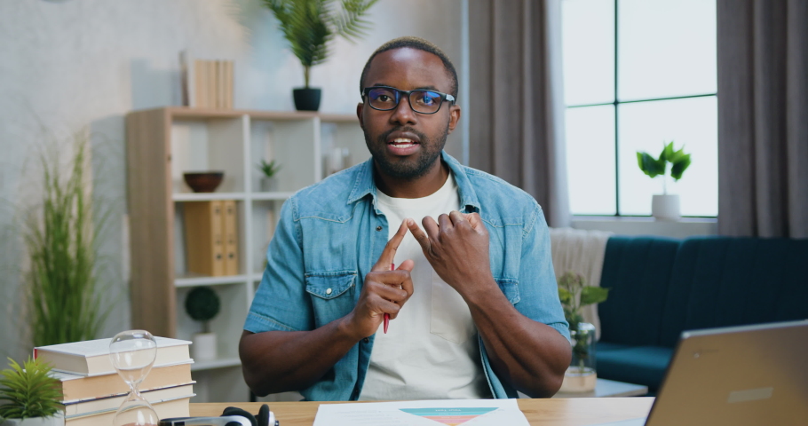 Good-looking positive confident skilled bearded african american in glasses sitting in front of camera at home and holding video conversation with client Royalty-Free Stock Footage #1083014881