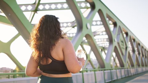 Rear view of young overweight woman in sports clothes walking outdoors in bridge.