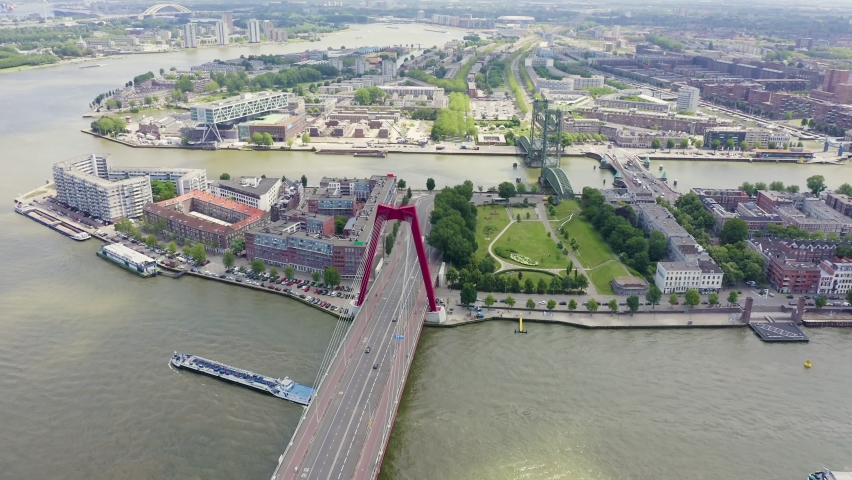 Rotterdam, Netherlands. The Willemsbrug cable-stayed bridge with distinctive red steel pylons and the historic De Hef vertical-lift railway bridge span the Nieuwe Maas river in a sweeping aerial hyperlapse. The Willemsbrug, opened in 1981, carries road traffic with bold red towers rising above the water, while nearby De Hef stands as a preserved industrial monument with green steel framework locked in a raised position. De Hef was the first vertical-lift bridge of this kind in Western Europe when completed in 1927, and has been a protected Rijksmonument since decommissioning in 1993. The flight path reveals the dense urban fabric of Rotterdam, with waterfront parks, historic harbor channels, and the modern skyline emerging in the distance under bright summer daylight. Hyperlapse
