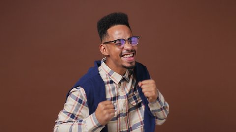 Excited jubilant young african american man 20s wears striped shirt glasses looks around doing winner gesture celebrate clenching fists say yes isolated on plain dark brown background studio portrait