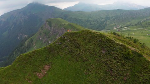 4K Drone flying fast around green hill in Italian Alps with woman standing on it wearing orange windproof jacket. Outdoor lifestyle, hiking, living in the wild, environment concept.