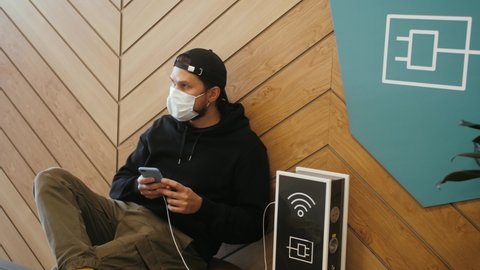 Person in medical mask uses a phone and charges it in a special place for charging with a usb wire. Man near the charging station in the mall charges the phone.