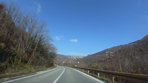Slow car driving on a curvy mountain road surrounded by trees. A mountain village in the distance. Autumn, blue clear sky