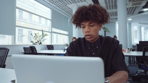 African American employee using laptop in office and mumbling something, diverse coworkers on background. Arc shot man working online, browsing internet. Concept of business