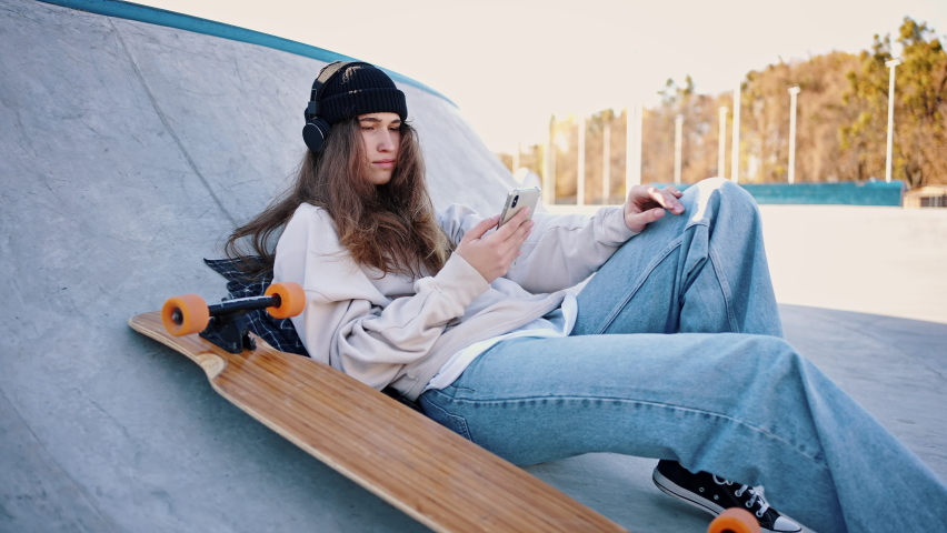 Casual dressed teenage girl lies on a springboard in a skatepark listening to music. Portrait of a girl and her longboard.