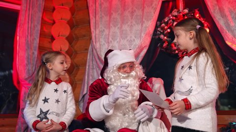 Children standing near Santa and talking to him. Kids sharing pictures and their Christmas dreams with Santa Claus. Santa asking girls about their wishes for holiday.