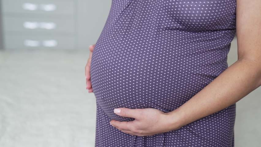 Expectant lady standing half-turned in her bedroom by the bed. Woman expecting a baby wearing a nightgown and stroking her belly. Close up.