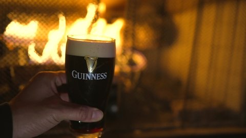 Chicago, IL USA - November 7 2021: This video shows an anonymous hand holding a guinness pint of beer in front of an inviting fireplace.