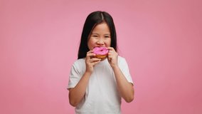Sweet Tooth. Korean Little Girl Eating Donut Smiling To Camera Posing Standing Over Pink Studio Background. Child Enjoying Doughnut Dessert. Unhealthy Sugary Food Concept. Slowmo