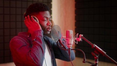 Music Production Concept. Young Black Guy In Professional Headphones Singing Song To Microphone At Sound Record Studio With Red Lighting, African American Male Singer Recording New Track, Slow Motion