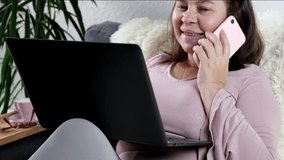 close-up of modern laptop and smartphone in female hands, middle-aged woman of 50 years old in pink sweater sits on sofa, reads news, communicates on Internet, concept of technology, stay at home