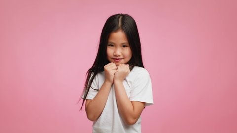 Shy Japanese Little Girl With Long Brunette Hair Posing Holding Fists Near Chin Smiling To Camera Standing Over Pink Studio Background. Portrait Of Cute Asian Preschool Child