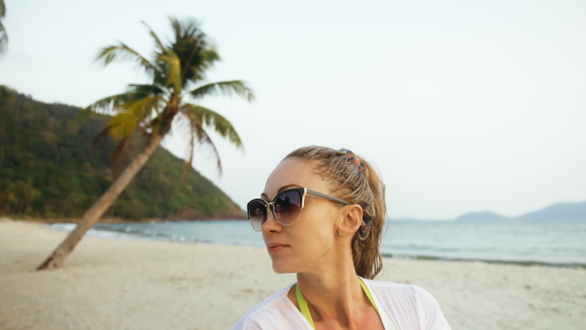 Close up portrait woman in a white tunic shirt on beach, near stormy sea, tilted palm tree. Girl in sunglasses. Female tourist walks