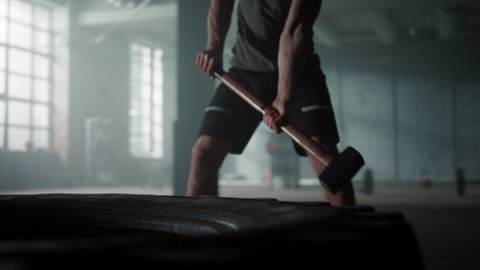 Unrecognizable athlete silhouette beating huge tire with sledgehammer. Closeup powerful man hitting hammer rubber wheel. Male professional bodybuilder training strength exercise at endurance workout 