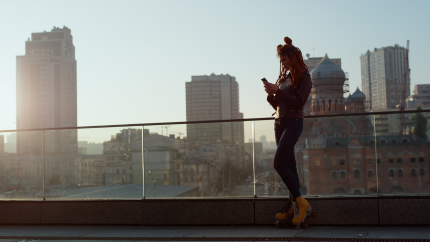 Side view of young woman silhouette in rollerblades staying on urban background. Stylish hipster girl using mobile phone outdoor. Beautiful female person looking to cellphone at rooftop.