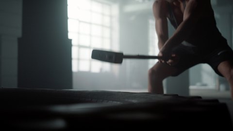 Closeup strong man hitting huge wheel with hammer in sport club. Professional bodybuilder silhouette exercise strength sledgehammer. Powerful athlete training workout using sports equipment in gym